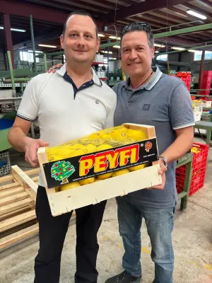 Zwei Männer stehen in einer Produktionshalle und halten gemeinsam eine Kiste mit frischem Obst – Symbol für enge Zusammenarbeit mit Erzeugern